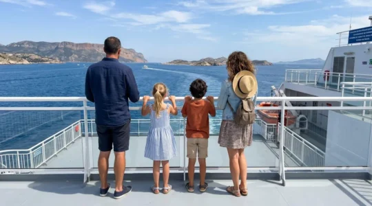 Une famille de quatre personnes observe l'horizon méditerranéen depuis le pont arrière d'un ferry moderne, photographiée de dos dans une ambiance détendue